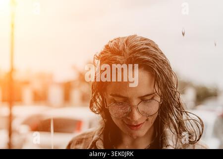 Femme lunettes pluie. Femme souriante portant des lunettes rondes marche à travers la pluie légère pendant l'heure dorée pour une ambiance urbaine paisible Banque D'Images