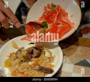 Gros plan des mains plaçant une salade de tomates fraîches et d'oignons, du riz, des légumes et de la viande sur une assiette sur un fond rustique, photographié en flou. Banque D'Images