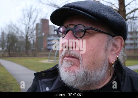 Homme senior avec une barbe grise et des lunettes rondes, portant une casquette plate et une veste noire. Plusieurs boucles d'oreilles cerceaux et une boucle d'oreille en croix ornent son oreille gauche. Le Banque D'Images