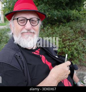 Un homme âgé est dehors, portant un chapeau rouge et des lunettes rondes noires. Il a une barbe blanche et est habillé d'une chemise noire avec un motif rouge en dessous Banque D'Images