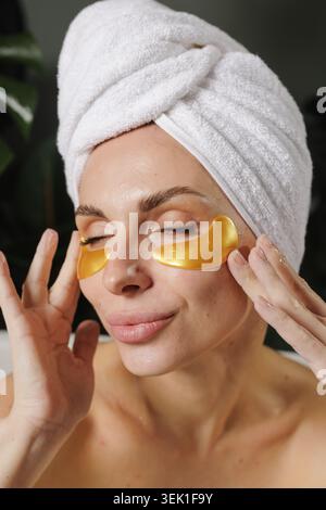 Une femme se détend tout en appliquant des patchs oculaires dorés dans un cadre de salle de bain serein, parfait pour les promotions de produits de spa ou de soins de la peau et les articles de bien-être. Banque D'Images