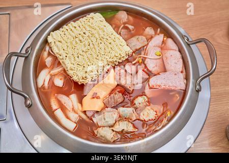 Budae-jjigae coréen dans un pot inoxydable, avec bouillon épicé chargé de saucisses, spam, fromage, boulettes et nouilles ramen bouillant activement. Banque D'Images