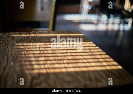 La lumière du soleil projette des ombres rayées sur une table de café en bois texturé. L'arrière-plan magnifiquement flou crée une atmosphère confortable et paisible l'après-midi. Banque D'Images
