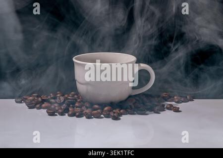 Un nuage de fumée entoure une tasse de café blanche avec des grains de café autour d'elle Banque D'Images