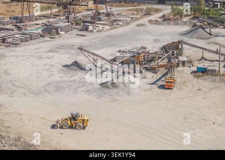 La vue aérienne capture un site de carrière industrielle occupé au Kirghizistan. La machinerie lourde fonctionne, y compris une chargeuse frontale jaune et un tombereau à benne orange, au milieu de tas de gravier et de sable. Banque D'Images