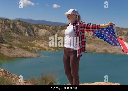 Femme se tient sur une colline tenant le drapeau américain, sentant un sentiment de liberté et de patriotisme au-dessus d'un lac turquoise serein entouré de collines sèches et Banque D'Images