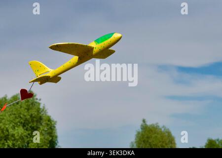 Plan à angle bas d'un avion de planeur jouet en mousse jaune volant haut dans le ciel bleu avec des nuages légers. L'avion jouet traîne une chaîne de petite patte rouge Banque D'Images
