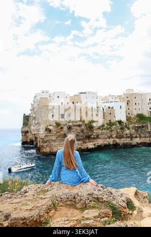 Voyageur féminin se relaxant sur la côte rocheuse avec de l'eau turquoise et ville balnéaire italienne historique en arrière-plan. Banque D'Images