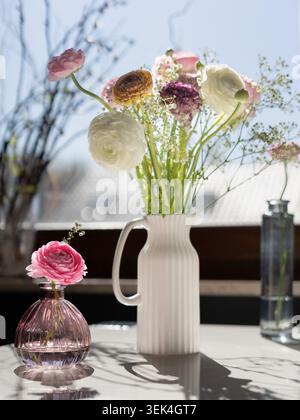 Composition florale brillante avec des fleurs de ranoncule disposées dans un vase blanc à côté d'une fenêtre ensoleillée Banque D'Images