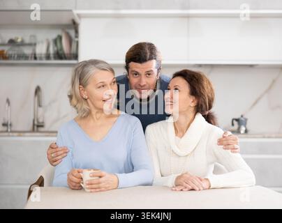 Mère âgée souriante et deux enfants adultes debout dans une cuisine lumineuse regardant la caméra Banque D'Images