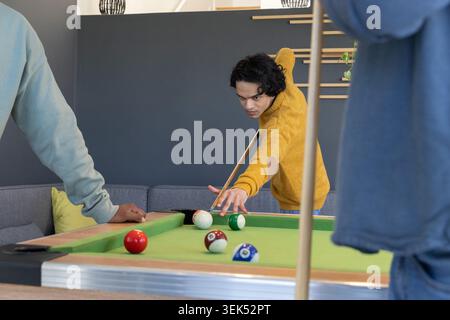 Homme asiatique portant un chandail moutarde et amis penchés sur une table de billard verte dans le salon, CUE de visée Banque D'Images