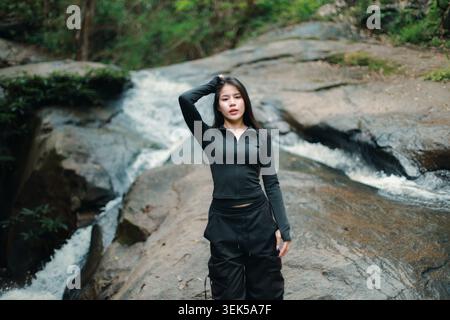 Belle jeune femme sereine debout sur un grand rocher à côté d'un ruisseau à écoulement rapide et cascade dans le paysage extérieur de la forêt verte nature se sentant calme Banque D'Images