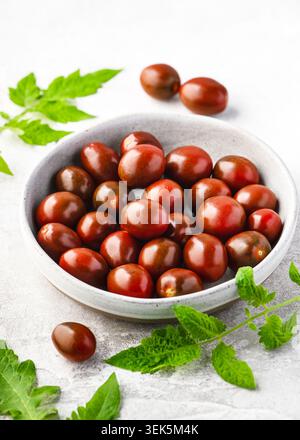 Mini tomates Kumato fraîches (Solanum lycopersicum) empilées dans un bol en céramique grise. Tomates cerises juteuses marron foncé avec des feuilles de tomate verte. Banque D'Images