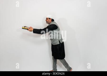 Homme indien portant des vêtements traditionnels et des lunettes, tenant un harmonium tout en regardant vers l'avant avec un léger sourire. Il est positionné contre un whi plaine Banque D'Images