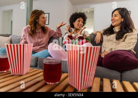 Diverses amies féminines vêtues de façon décontractée discutant sur le canapé dans le salon avec pop-corn et télécommande Banque D'Images
