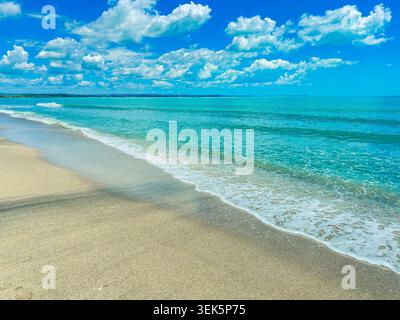 La lumière du soleil brille sur une plage avec du sable doux et des vagues claires qui roulent de l'océan. Les nuages dérivent à travers le ciel bleu au-dessus tandis que l'eau coule Banque D'Images