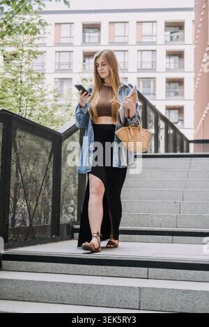 Jeune femme se tient sur les escaliers de la ville regardant smartphone tout en tenant un ordinateur portable. Prise de décision à la volée, vie professionnelle active, affaires Banque D'Images