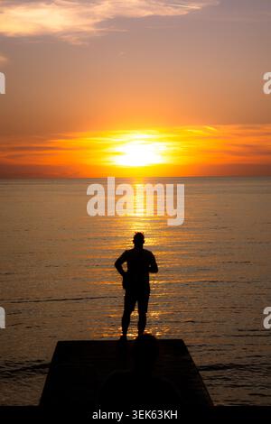 Silhouette d'une personne debout sur une jetée et regardant un coucher de soleil doré sur l'eau calme à la mer Banque D'Images