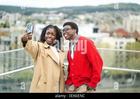 Heureux couple afro-américain marié voyageurs posant à un point de vue élevé dans la ville prendre selfie sur smartphone, souriant noir hommes et femmes partenaires touristes prendre des photos ensemble sur téléphone portable. Banque D'Images