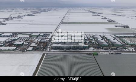 Vue aérienne de bâtiments industriels et de serres entourés de champs enneigés et d'éoliennes sous un ciel couvert à Middenmeer, North Hol Banque D'Images