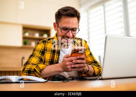 Jeune homme heureux utilise un ordinateur portable et un smartphone tout en travaillant à la maison. Banque D'Images