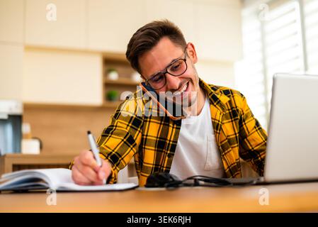 Travaillant à domicile, un jeune homme écrit des idées et des rappels, combinant la recherche en ligne avec la prise de notes traditionnelle. Banque D'Images