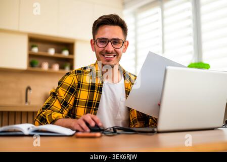 Avec une expression joyeuse, un homme utilise son ordinateur portable, semblant analyser des documents ou résoudre un problème en ligne. Banque D'Images