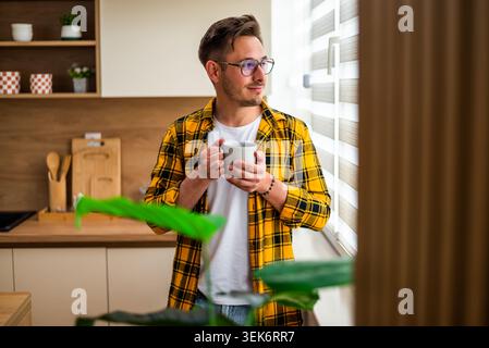 Le jeune homme se tient près de la fenêtre dans sa cuisine et boit du café. Il regarde dehors. Banque D'Images