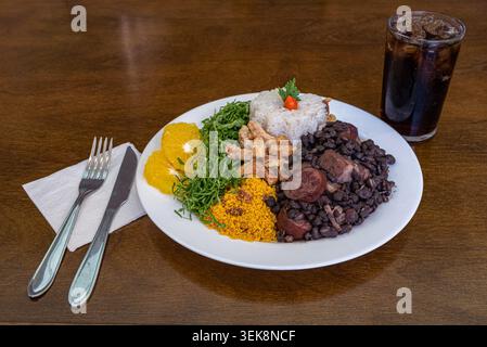 Feijoada traditionnelle sur assiette blanche avec haricots noirs, riz et oranges. Table rustique avec argenterie et boisson offrant une ambiance chaude saturée Banque D'Images
