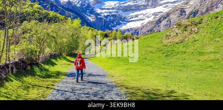 Randonneur portant une veste rouge marche sur un sentier de gravier dans les Pyrénées françaises, entouré de montagnes enneigées et de paysages verdoyants Banque D'Images