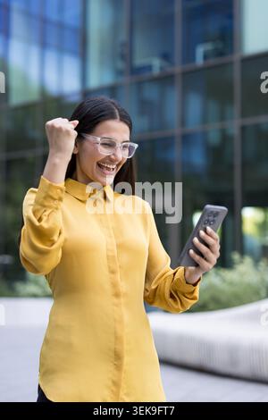 Jeune femme souriante levant le poing en célébration tout en regardant son smartphone à l'extérieur d'un immeuble de bureaux en verre bleu, montrant l'excitation et la réussite Banque D'Images