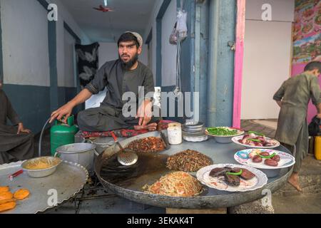 Vendeur de rue afghan portant une robe traditionnelle préparant divers plats cuisinés sur une grande plaque de métal. Banque D'Images