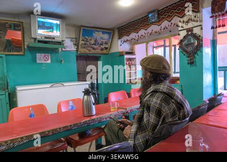 Homme dans un chapeau de pakol et chemise à carreaux buvant du thé et regardant la télévision dans un salon de thé traditionnel. Banque D'Images