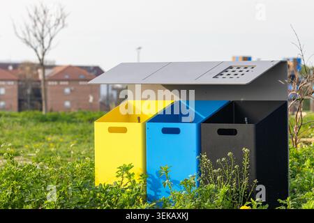 Trois bacs de recyclage colorés en jaune, bleu et noir se dressent à l’extérieur pour le tri et le recyclage des déchets à proximité d’un quartier résidentiel, entouré par Banque D'Images