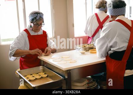 Femme hispanique âgée préparant de petites tartes dans une boulangerie commerciale lumineuse Banque D'Images
