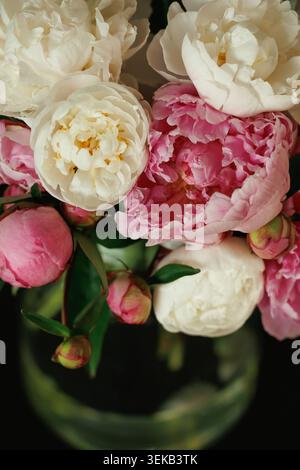 Bouquet de pivoines blanches et roses mélangées dans un vase en verre transparent, boutons et fleurs ouvertes, fond sombre Banque D'Images