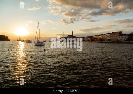 Coucher de soleil sur la ville côtière de Rovinj avec des voiliers amarrés dans le port et une tour d'église sur la colline, Croatie. Banque D'Images