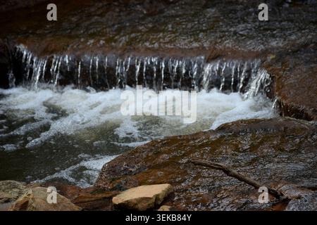 De l'eau claire tombant des rochers dans un ruisseau voisin à côté d'un sentier forestier populaire. De grandes roches brunes, scintillantes avec de l'eau de source, tapissent ses côtés. Banque D'Images