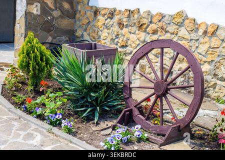 Un lit de jardin décoratif avec une vieille roue de wagon en bois et de petites plantes contre un mur de pierre. décoration de jardin, style rustique, aménagement paysager. Banque D'Images