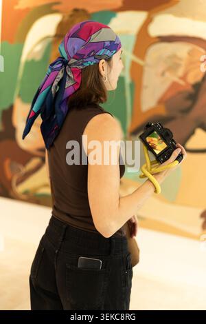 Vue de profil nette et détaillée d'une femme portant un foulard coloré et tenant un appareil photo numérique. Elle se tient devant un grand tableau abstrait Banque D'Images