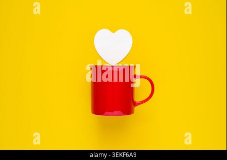 Un mug rouge vif avec une note blanche en forme de coeur sur un fond jaune vif. Banque D'Images