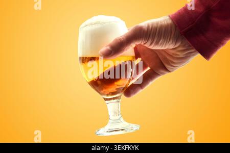 Main tenant un verre à bière givré avec une mousse épaisse sur un fond jaune chaud, soulignant le rafraîchissement, les loisirs et l'atmosphère de boisson sociale. Banque D'Images