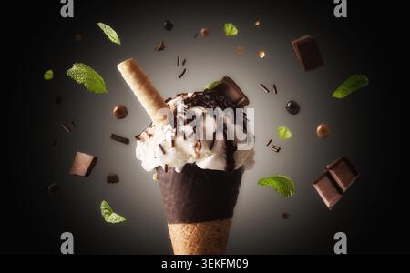 Crème glacée dans un cornet de gaufre enrobé de chocolat surmonté de saupoudrées de chocolat, de morceaux de chocolat et d'un bâtonnet de gaufrette, entouré d'ingrédients flottants Banque D'Images
