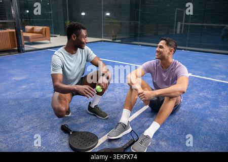 Divers amis masculins dans des vêtements de sport discutant sur un court de padel bleu tenant une balle jaune et deux raquettes. Amitié, occasionnel, athlétique, récréatif, verre, installation, canapé Banque D'Images