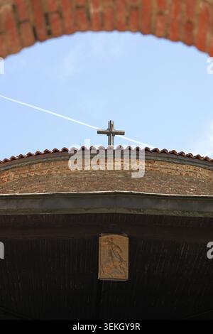 Ancien bâtiment religieux avec une croix sur son toit en tuiles, vu à travers une arche en briques, avec un bas relief religieux classique ornant l'avant-toit en bois Banque D'Images