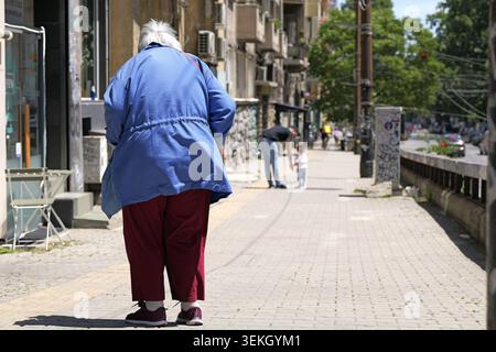 Femme âgée en veste bleue et pantalon rouge marchant le long d'un trottoir de la ville, vue arrière pleine longueur avec piétons flous et circulation, stre urbain ensoleillé Banque D'Images