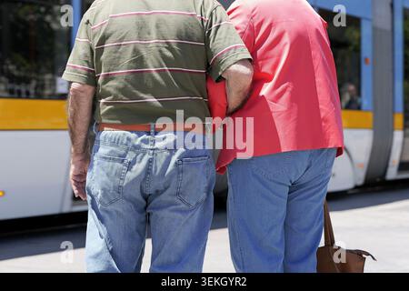 Couple de personnes âgées marchant bras dans bras, symbolisant l'amour, l'unité et le soutien dans leur mode de vie senior en attendant les transports en commun dans une ville Banque D'Images