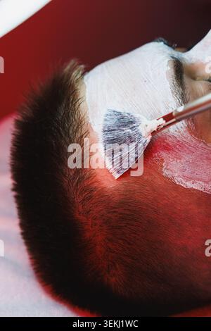 Le client reçoit une thérapie de lumière rouge lors d'un rendez-vous de cosmétologie. Un thérapeute applique un produit facial à l'aide d'un pinceau. L'environnement a une focalisation Banque D'Images