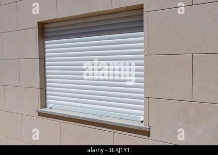Un mur extérieur contemporain avec de grands carreaux de pierre beige et une fenêtre protégée par un système de volets roulants blancs Banque D'Images