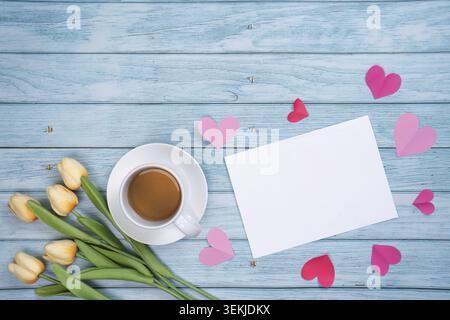 Une scène chaleureuse avec une tasse de café, des tulipes jaunes et une carte vierge entourée de coeurs roses sur un fond de bois, parfait pour la fête des mères Banque D'Images